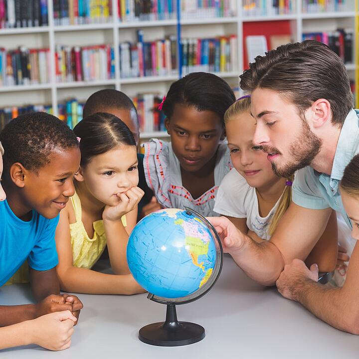 Eine multikulturelle Schulklasse ist in einer Bücherei um einen kleinen Globus versammelt.